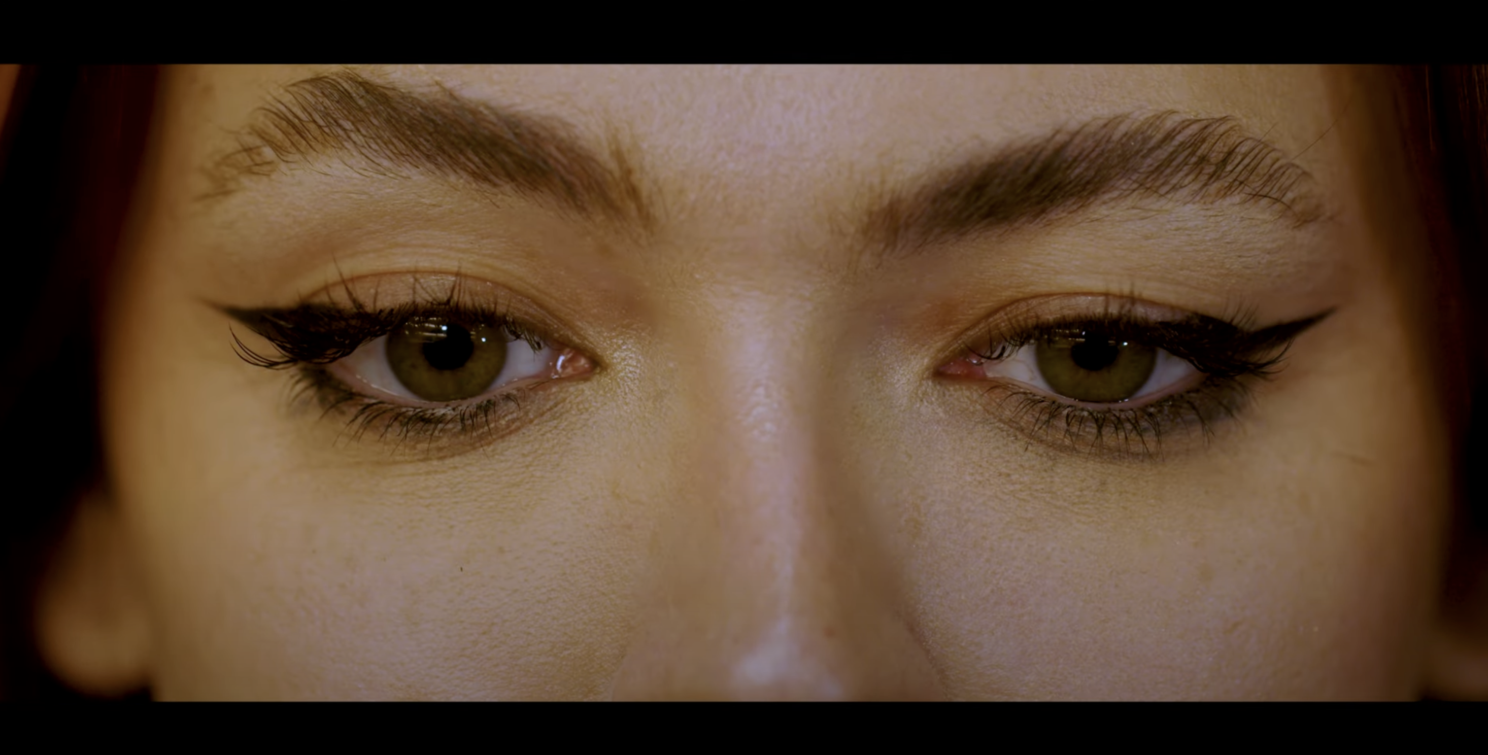 A close up image of a woman's face from the nose up. She is wearing black eyeliner and a downcast expression.